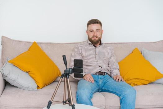 Smiling bearded guy filming video using smartphone on tripod online while photo