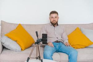 Smiling bearded guy filming video using smartphone on tripod online while photo
