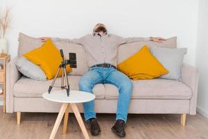 hombre barbudo sonriente filmando video usando un teléfono inteligente en un trípode en línea mientras foto