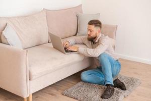 Young attractive man sitting on sofa at home, working on laptop online, using photo