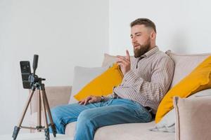 Smiling bearded guy filming video using smartphone on tripod online while photo