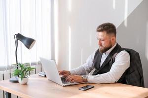 Caucasian bearded business man is sitting at window table using his laptop photo