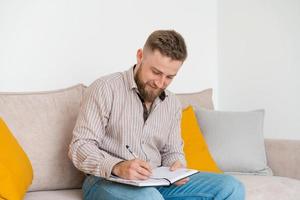 Successful bearded young man writes notes in notebook while sitting at home photo