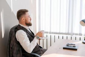 disfrutando de un café recién hecho en el trabajo. confiado joven barbudo en ropa de negocios foto