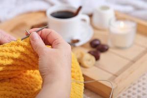 hands of a woman knitting sweater with yellow soft wool. coffee and snacks on a wooden tray on sofa. cozy ambient weekend morning. photo