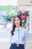 Beautiful high school Asian student girl in school uniform stands and smiles happily with braces on her teeth while she confidently while she looks at the camera with building in city as a background photo