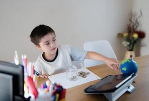 School kid pointing finger at world map and putting money coins into clear jar, Child saving money for travel, Children learning about saving for education and future concept photo