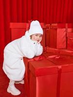 Cute little girl in white coat by the huge red christmas presents boxes on red background. Holidays, shopping, fun, fashion photo