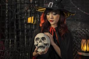 Chica asiática disfrazada de bruja sosteniendo un cráneo humano para el concepto de truco o trato de la fiesta de Halloween con fondo negro oscuro con espacio para copiar foto