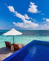 Beautiful luxury water villas resort, infinity swimming pool, beach chairs. Tranquil sunny sky, ocean lagoon beach background. Couple summer vacation holiday travel. Paradise island, travel landscape photo