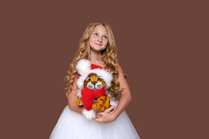 Child plays with cute little Christmas tiger toy in white dress on brown photo