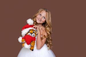 Child plays with cute little Christmas tiger toy in white dress on brown photo