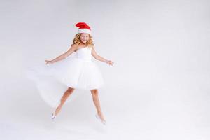 Joyful pretty girl in Red Santa Claus Hat and white dress Laughs and jumps photo