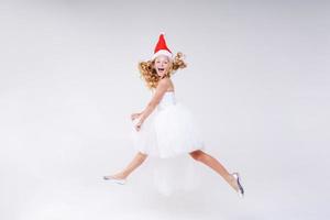 Little joyful girl in red Santa hat and white beautiful dress jumps from photo