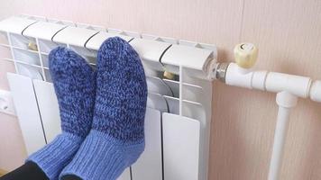 una mujer se calienta los pies junto al radiador en un frío día de invierno con calcetines azules de lana. sistema de calefacción central. elevados costes de calefacción durante la estación fría. video