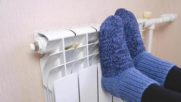 una mujer se calienta los pies junto al radiador en un frío día de invierno con calcetines azules de lana. sistema de calefacción central. elevados costes de calefacción durante la estación fría. video