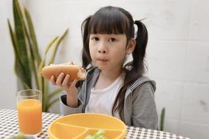 joven asiática desayunando y jugo de naranja en la mesa en casa todavía feliz. alimentación saludable, comida y bocadillos, sándwich de jamón y queso, niño feliz comiendo foto