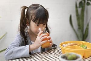 hermosa joven bebiendo jugo de naranja y desayunando en la mesa en casa. niño feliz comiendo, jugo de frutas, alimentación saludable, comida y bocadillos foto