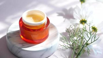 Skincare product in orange container on marble platform with flower beside video