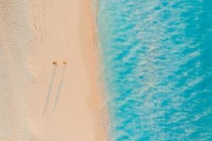 Aerial view of couple walking on beach with sunlight close to turquoise sea waves. Top view of summer beach landscape, romantic inspirational couple vacation, romance holiday. Freedom travel template photo
