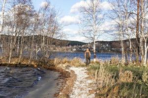 young man in hood from back walking on shore of lake among birches photo