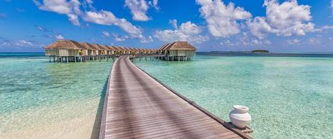 Sunny Maldives island, luxury water villas resort and wooden pier. Beautiful sky and clouds and beach background for summer vacation holiday and travel concept. Amazing panoramic beach view, tourism photo