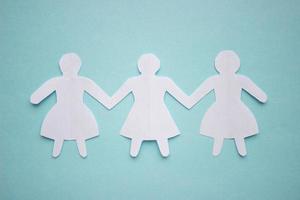 Figures of three girls in dresses holding hands, cut out of white paper. In the center of the photo on a blue background