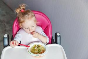 Infant baby with dirty face eating soup herself with spoon. Cute little girl sitting in high baby chair in kitchen at home. Maternity family childhood concept. photo