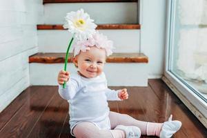Little smiling baby girl one year old wearing spring wreath siting on floor in bright light living room near window and playing with gerbera flowers. Happy kid playing at home. Childhood concept. photo