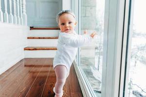 Little crawling baby girl one year old siting on floor in bright light living room near window smiling and laughing photo