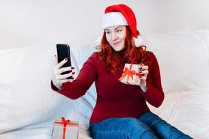 una joven pelirroja con sombrero de santa se sienta en el sofá de casa y usa el teléfono móvil para chatear por video. saludo en línea con año nuevo y navidad. foto