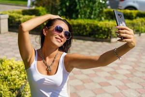 una joven alegre se toma selfie de las manos con el teléfono mientras se sienta en el parque foto