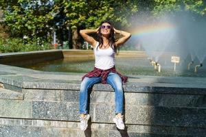 Cheerful young woman in sunglasses sits against backdrop fountain with rainbow photo