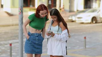 dos mujeres jóvenes miran juntas el teléfono mientras están afuera cerca de una calle video