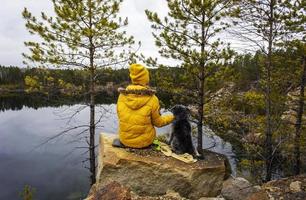 Young woman in yellow clothes from the back with a fluffy gray dog sitting on a rocky lake near pine trees. Traveling with pets concept photo