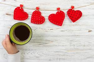 Above view of female hand holding hot cup of coffee with red heart on wood table. Photo in vintage color image style
