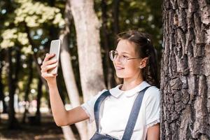 A schoolgirl in glasses takes a selfie on a smartphone near a tree in the park. photo