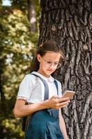 A schoolgirl in glasses stands near a tree and looks into a smartphone in the park. Vertical view photo