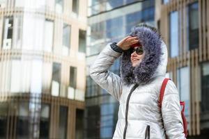 Happy young woman in winter jacket with backpack near building photo