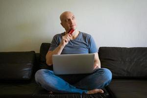 Emotional bald man on sofa with laptop and credit card in hand photo