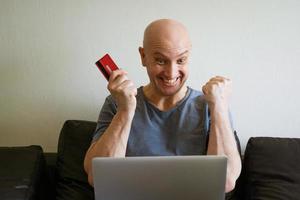 hombre calvo emocional en el sofá con computadora portátil y tarjeta de crédito en la mano foto