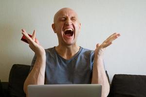 Emotional bald man on sofa with laptop and credit card in hand photo