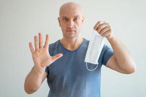 A bald man holds a medical mask in his hands, his other hand held out in protest photo