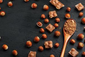 Chocolate with hazelnuts and a wooden spoon with cocoa on a dark background, surrounded by nuts in the shell and peeled.