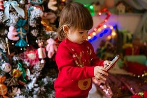 Little girl in red sweater is watching cartoons on phone next to christmas tree photo