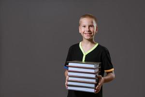 niño alegre sosteniendo una pila de libros sobre un fondo gris foto