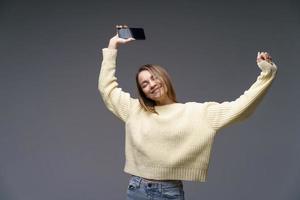 mujer joven en suéter amarillo sobre fondo gris bailando con el teléfono en la mano foto