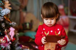 Little girl in red sweater is watching cartoons on phone next to christmas tree photo