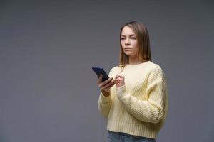 young woman writes sms on the phone in a yellow sweater on a gray background photo