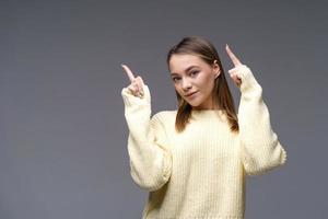 Cheerful young woman showing thumbs up in yellow sweater on gray background photo